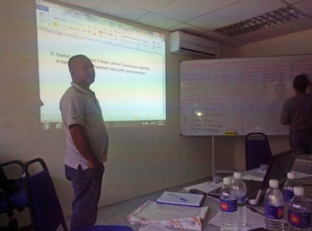 Berjaya in a classroom in front of a screen on to which information is projected.