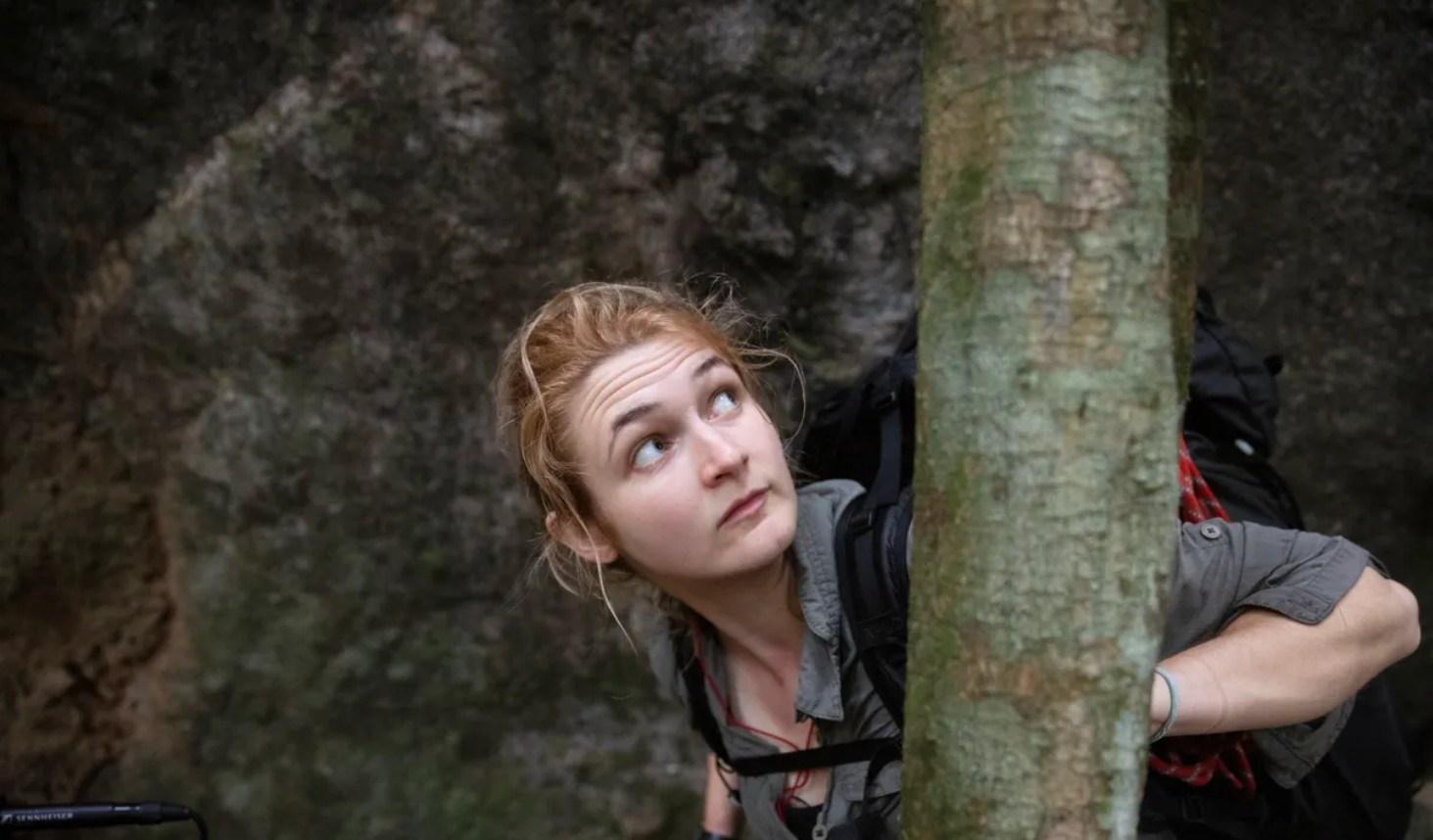 Lucy Shepherd peers around a tree trunk in a forest